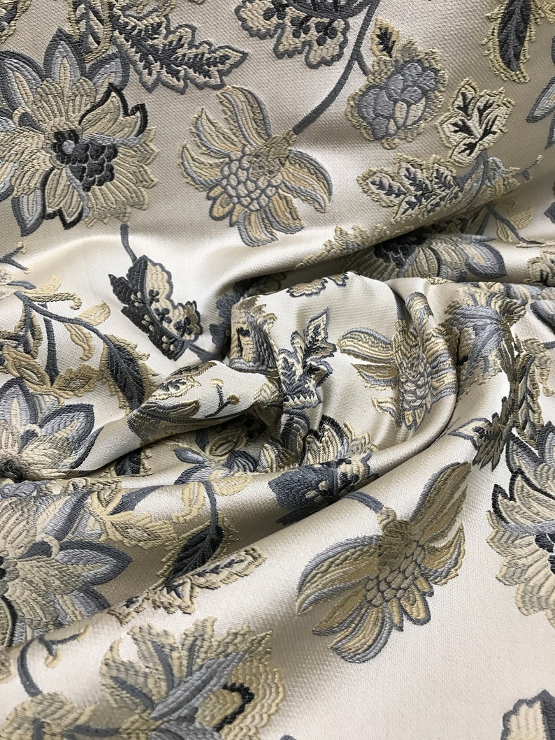 May include: Close-up of a white fabric with a floral pattern. The fabric has a raised texture and features gray and beige flowers.