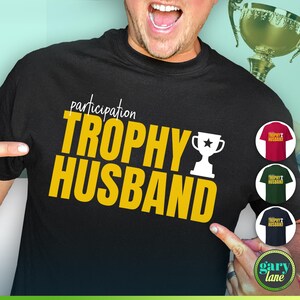 May include: A black t-shirt with the text "participation trophy husband" in yellow letters. A white trophy icon is next to the text. The shirt is being modeled by a person.
