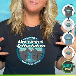 May include: A black t-shirt with a blue and white graphic design featuring a mountain scene and the text "PLEASE STICK TO THE RIVERS & THE LAKES THAT YOU'RE USED TO".