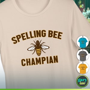 May include: A beige t-shirt with brown text that reads "Spelling Bee Champian" and a cartoon bee illustration. The t-shirt is shown on a hanger with three other t-shirts in different colors.