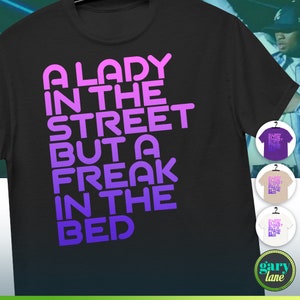 Può includere: Una maglietta nera con un design sfumato viola e rosa. Il testo sulla maglietta recita "A Lady in the Street But a Freak in the Bed."