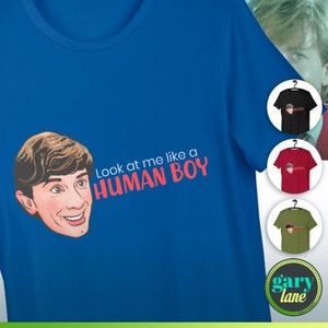 Puede incluir: Una camiseta azul con una ilustración de dibujos animados de una cara y el texto "Look at me like a HUMAN BOY".