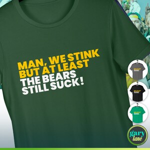 May include: Green t-shirt with the text "MAN, WE STINK BUT AT LEAST THE BEARS STILL SUCK!" in yellow and white. The shirt is displayed with three other color options: gray, black, and green. The brand logo is visible at the bottom.