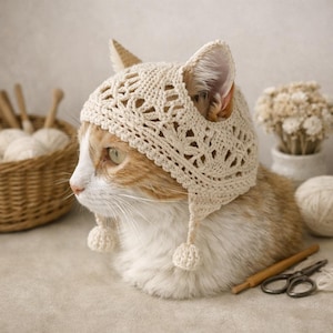 May include: A cream-colored crocheted cat hat featuring an intricate openwork pattern, ear cutouts, and two dangling pom-poms at the chin. The headwear is crafted from fine yarn and designed to fit over the ears of a cat, providing a snug appearance.