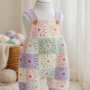May include: A pastel-colored crochet baby romper with a floral granny square design. The overalls feature a mix of pink, purple, green, and cream squares, with wooden buttons on the straps. The garment is displayed on a white mannequin.