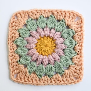 May include: A square crochet piece with a floral design. The centre is yellow, surrounded by pink petals, green leaves, and a peach border. The yarn has a soft texture, suitable for various craft projects. The dimensions are approximately 20 cm x 20 cm.