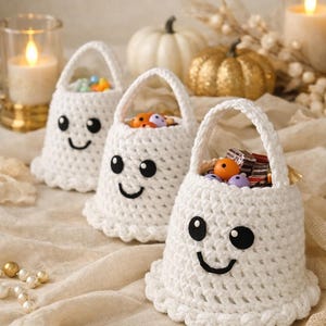 May include: Three white crochet ghost-shaped baskets with black eyes and smiles, filled with colorful candies. The baskets have handles and scalloped edges. Decorative pumpkins and candles are in the background, creating a festive Halloween theme.