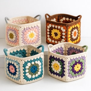 May include: Four square crochet baskets in various colors. Each basket features a floral design in the center, with a neutral border and handles. Colors include pink, brown, blue, and purple. These decorative baskets are ideal for storage.