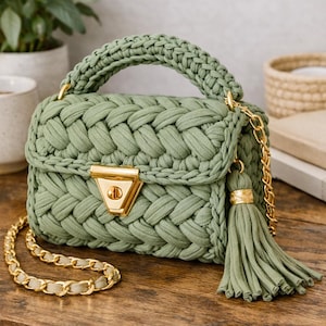 May include: A sage green crochet handbag featuring a thick braided texture, a gold-tone triangular twist lock closure, a matching top handle, and a long gold chain strap. A decorative sage green fabric tassel hangs from the side of the bag.