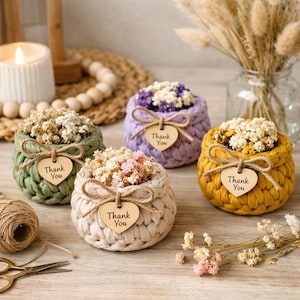 Può includere: Una collezione di cinque piccoli cestini all'uncinetto nei toni del verde, viola, giallo e crema, ciascuno riempito di fiori secchi. Ogni cestino ha un'etichetta di legno a forma di cuore con la scritta "Thank You".