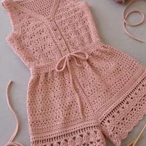 May include: A sleeveless, light pink crochet romper with a V-neck and wooden buttons. The romper features a tie at the waist and decorative edging on the shorts. A ball of yarn and crochet needles are also visible.