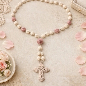 May include: A handmade rosary featuring cream-colored and dusty pink crocheted beads linked by silver-toned metal connectors, terminating in a detailed, openwork crocheted cross pendant with a matching cream-colored yarn finish.