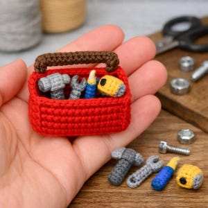 Puede incluir: Una pequeña caja de herramientas roja de ganchillo con un asa marrón, que contiene pequeños martillos grises, llaves y herramientas amarillas y azules. Se colocan herramientas adicionales junto a la caja de herramientas. Las herramientas están hechas a mano.