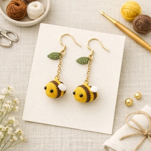 May include: A pair of handmade crochet bee earrings. The earrings feature yellow and brown striped bee bodies with white wings, green leaf accents, and gold-colored hooks. The earrings are displayed on a white card.