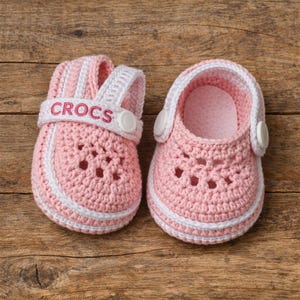 Puede incluir: Un par de zapatos de bebé tejidos en ganchillo de color rosa claro con ribete blanco y una tira trasera blanca. El zapato izquierdo tiene la palabra CROCS bordada en hilo rosa en la tira. Ambos zapatos tienen un diseño perforado y botones.