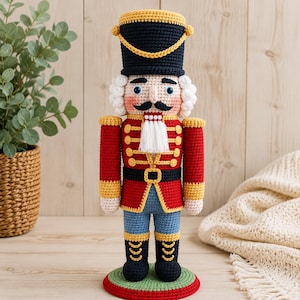 May include: A crocheted nutcracker doll featuring a tall black hat with yellow trim, a red jacket with yellow accents, blue trousers, and black boots. The figure has white hair, a black moustache, and stands on a round green and red base.