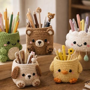 Può includere: Cinque portapenne all'uncinetto a forma di animali e una nuvola. La collezione include un dinosauro verde, un orso marrone, una nuvola bianca con gocce di pioggia pendenti, un cucciolo beige con orecchie cadenti e un pulcino giallo, pieni di penne.