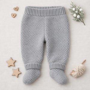 May include: A pair of gray, crocheted baby pants with attached feet. The pants have a ribbed waistband and cuffs. Wooden star and heart-shaped decorations are scattered around the pants, along with a small bundle of yarn and a sprig of white flowers.