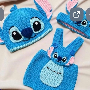 May include: A set of handmade blue crochet baby clothes, including a hat, headband, and overalls, all featuring a cartoon character design with pink and black accents. The hat has large ears and a character face.