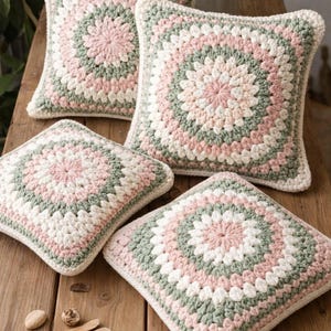 May include: Four square crochet pillows in shades of pink, white, and green arranged on a wooden surface. The pillows have a circular pattern in the center and a solid border. The pillows are a decorative home decor item.