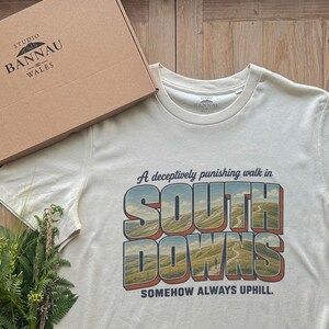 May include: A cream-colored short-sleeve t-shirt featuring a graphic of rolling hills with the text A deceptively punishing walk in SOUTH DOWNS SOMEHOW ALWAYS UPHILL. The shirt is accompanied by a cardboard box labeled STUDIO BANNAU WALES.
