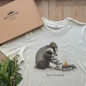 Puede incluir: Una camiseta de manga corta color hueso con un gráfico de un tejón con gorro verde, vertiendo líquido de una tetera en una taza junto a una fogata. El texto "Brew Secured." está impreso debajo del gráfico. Se ve una caja de cartón con la etiqueta "STUDIO BANNAU WALES".
