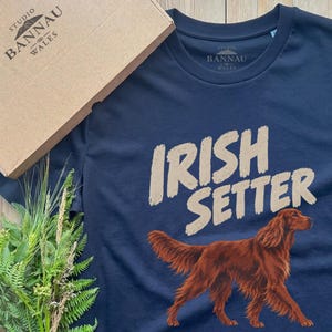 May include: A navy blue short-sleeved t-shirt featuring a graphic of a reddish-brown Irish Setter dog in profile. The text "IRISH SETTER" is printed in a light beige color above the dog. The shirt includes a branded inner neck label.