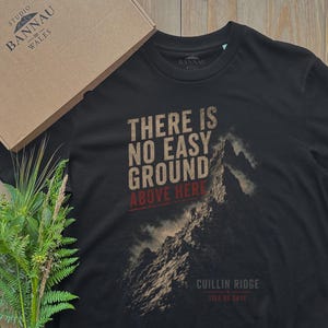 May include: A black short-sleeved t-shirt featuring a graphic of a mountain peak with the text "THERE IS NO EASY GROUND ABOVE HERE" in tan and red, and "CUILLIN RIDGE ISLE OF SKYE" in white. The shirt is accompanied by a cardboard box and green ferns.