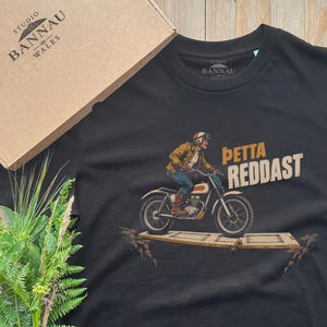 Può includere: Una t-shirt nera a maniche corte con la grafica di una persona in sella a una motocicletta su una tavola di legno. Il testo PETTA REDDAST è stampato in oro accanto al guidatore. La maglietta include un'etichetta sul collo e una scatola.