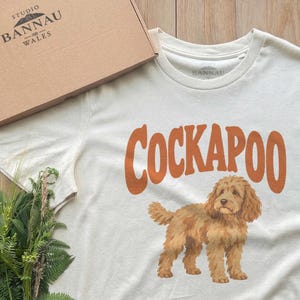 Peut inclure: Un t-shirt à manches courtes blanc cassé avec le mot COCKAPOO en grandes lettres orange au-dessus d'une illustration graphique d'un chien Cockapoo brun doré. Le t-shirt est présenté avec une boîte en carton marquée Studio Bannau Wales.