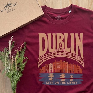 Può includere: Una t-shirt a maniche corte color bordeaux con una stampa grafica della parola DUBLIN in lettere beige sopra un ponte stilizzato, una fila di edifici e il testo CITY ON THE LIFFEY in blu, con una scatola di cartone etichettata STUDIO BANNAU WALES.