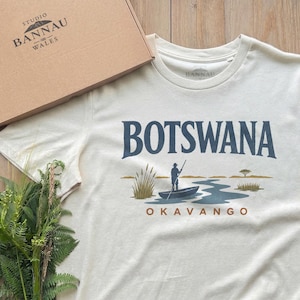 May include: A cream-colored short-sleeve t-shirt featuring a graphic print with the text "BOTSWANA" in dark blue and "OKAVANGO" in orange. The illustration depicts a person in a small boat on a river with reeds and a tree in the background.