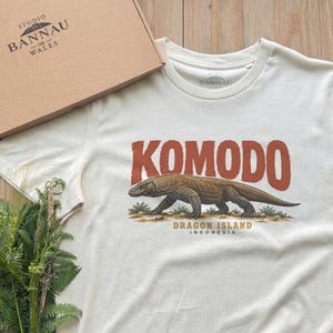 Peut inclure: Un t-shirt à manches courtes de couleur crème avec une illustration de dragon de Komodo. Le texte KOMODO est imprimé en grandes lettres orange au-dessus du lézard, avec les mots DRAGON ISLAND INDONESIA en plus petits caractères en dessous.