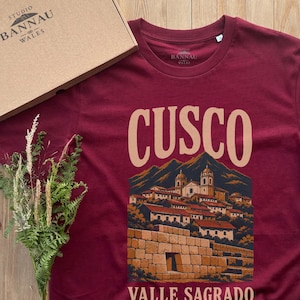 Peut inclure: Un t-shirt bordeaux à manches courtes avec une impression graphique de la ville de Cusco et des montagnes des Andes. Le texte CUSCO est imprimé en grandes lettres beiges au-dessus de l'illustration, avec VALLE SAGRADO en dessous.