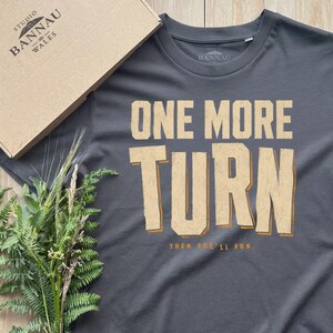 Puede incluir: Una camiseta de manga corta gris carbón con el texto ONE MORE TURN en letras beige grandes y desgastadas, con el texto más pequeño THEN SHE'LL RUN debajo. La camiseta se muestra junto a una caja de cartón y helechos verdes.