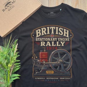 May include: A black t-shirt featuring a graphic of a red and black stationary engine with a large flywheel. The text reads BRITISH STATIONARY ENGINE RALLY 1974, FLYWHEELS, RESTORATION, SHOW FIELD, and BRITAIN. The shirt is paired with a cardboard box.