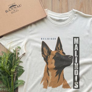 Peut inclure: Un t-shirt à manches courtes blanc cassé avec une illustration de tête de chien Malinois belge. Le texte "BELGISCH" est inscrit au-dessus du chien, et "MALINOIS" est imprimé verticalement dans un bloc rectangulaire noir à droite.