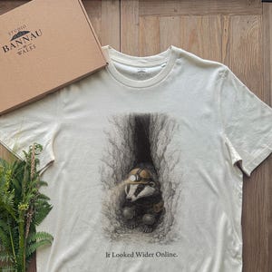 Puede incluir: Una camiseta de manga corta color hueso con una ilustración detallada de un tejón con casco de minero dentro de un túnel estrecho. Debajo del gráfico, el texto dice: "It Looked Wider Online." La camiseta se presenta junto a una caja de cartón.