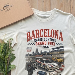 May include: An off-white short-sleeve t-shirt featuring a graphic print of a Barcelona Radio Control Grand Prix 1986 race scene. The design includes a remote control transmitter, a race car, a battery pack, and tools, with text detailing the event.