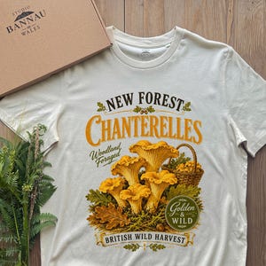 May include: An off-white short-sleeve t-shirt featuring a graphic of yellow chanterelle mushrooms in a basket with the text New Forest Chanterelles, Woodland Foraged, Golden & Wild, and British Wild Harvest, accompanied by a brown cardboard box.