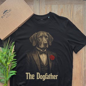Op de afbeelding: Een zwart t-shirt met een afbeelding van een hond in een smoking met een rode roos op de revers, een vlinderdas en de tekst "The Dogfather" eronder gedrukt. Het t-shirt wordt vergezeld door een kartonnen doos met "Studio Bannau Wales" en een varen.