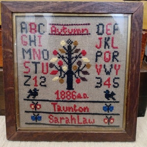 以下が含まれることがあります： 木製フレームに入ったクロスステッチのサンプル。アルファベット、数字、カラフルな葉の木が特徴です。「Autumn」の文字が上部にステッチされ、1886年と「Sarah Law」の名前が下部にあります。