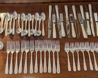 May include: A large collection of silver-toned flatware featuring ornate floral embossed handles. The set includes various sizes of forks, spoons, and knives, along with a large serving ladle and several small demitasse spoons arranged in rows.