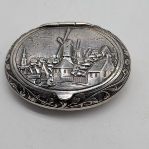 May include: Oval silver-toned box with a detailed relief scene of a village with windmills, houses, and a church. The box has a hinged lid and ornate scrollwork around the edges, showcasing a vintage aesthetic.