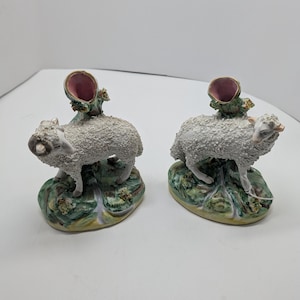 May include: Two ceramic ram figurines. Each ram has a textured, white wool coat, brown horns, and is standing on a green and yellow base. A pink-lined, vase-like structure is on the ram's back.