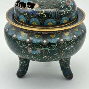 May include: A dark green, three-legged cloisonné incense burner with a lid. The burner features floral and vine designs in blue, yellow, and white, with gold trim. The lid has a decorative opening.