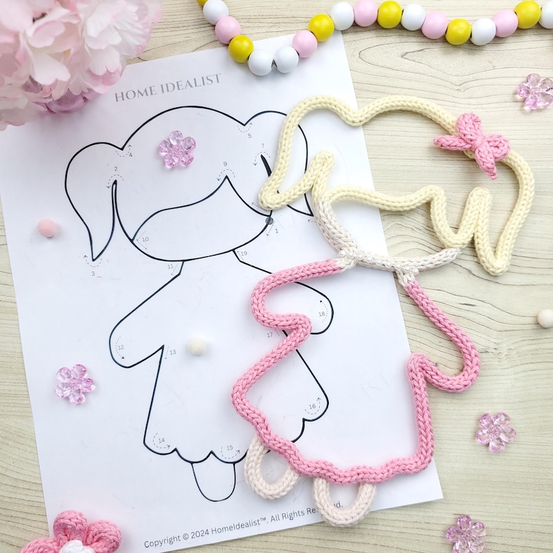 May include: A color-by-number illustration of a girl with a pink and cream-colored crocheted outline. The image includes the text "HOME IDEALIST" and copyright information. Decorative elements include pink flowers and wooden beads.