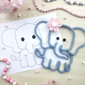 May include: A light blue knitted elephant with a pink flower on its head rests on a wooden surface next to a paper elephant outline. Pink and white wooden beads and small pink pom-poms are scattered around the elephant.