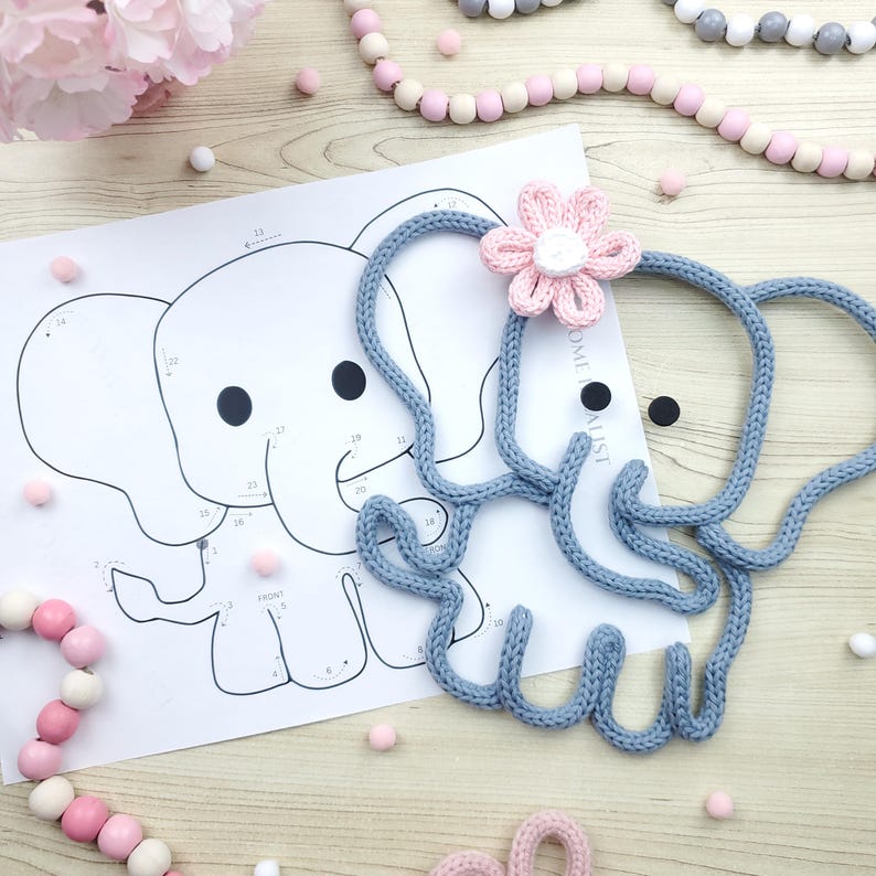 May include: A light blue knitted elephant with a pink flower on its head rests on a wooden surface next to a paper elephant outline. Pink and white wooden beads and small pink pom-poms are scattered around the elephant.