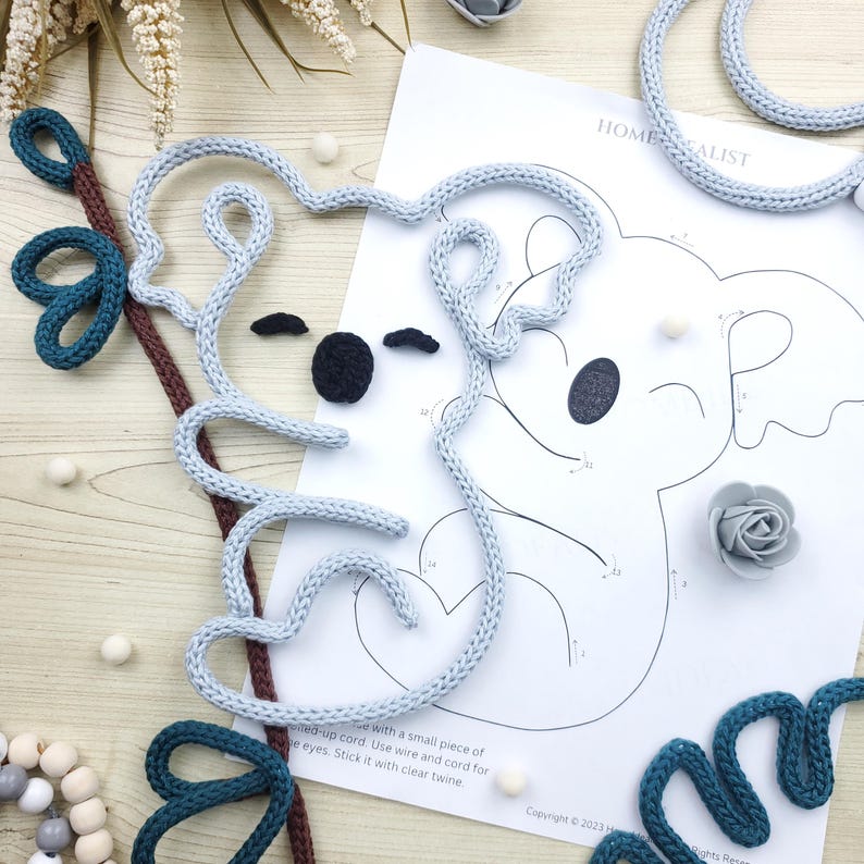 May include: A light blue knitted koala outline with black eyes and a nose, along with a printed koala illustration. Additional elements include teal and brown knitted accents, small white balls, and a gray rose. The image includes the text "HOME ALIST".